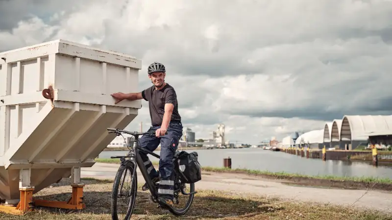 JobRadler in Arbeitshose lehnt an Container, hinter ihm Wasser JobRadler in Arbeitshose lehnt an Container, hinter ihm Wasser