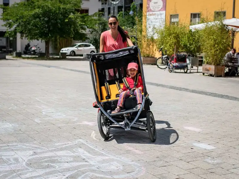 Eine Mutter fährt das Chike e-Bike in der Stadt. Ihr Kind sitzt angeschnallt darin. Beide lachen Eine Mutter fährt das Chike e-Bike in der Stadt. Ihr Kind sitzt angeschnallt darin. Beide lachen