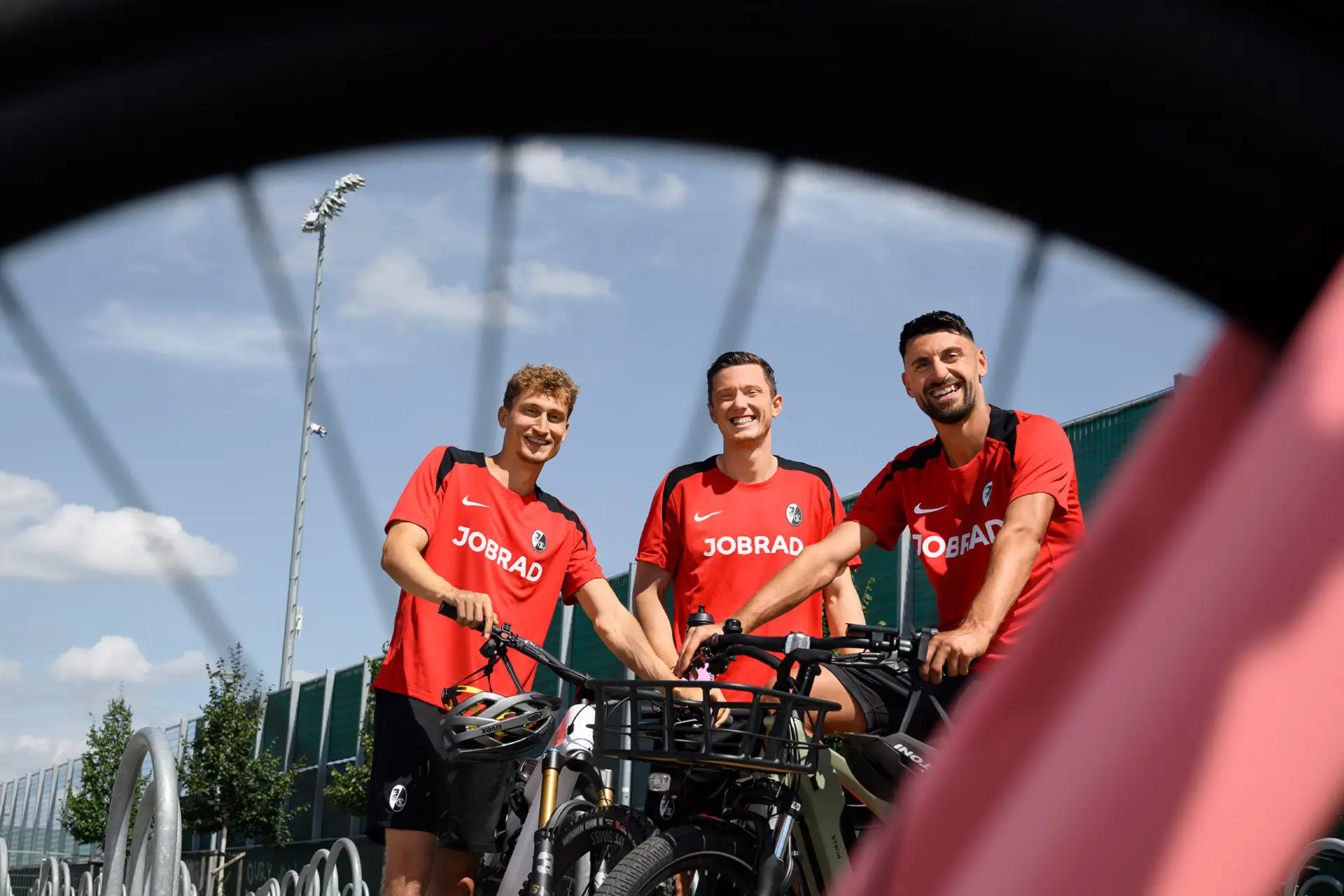 3 SC Spieler stehen mit ihren Fahrrädern vor dem Stadion und grinsen in die Kamera